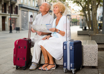 Fototapeta na wymiar Traveling glad cheerful positive smiling mature spouses resting on stone bench on city street, looking map