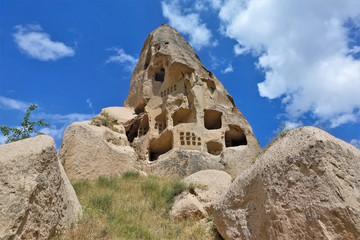 Ancient conical rock of Cappadocia against blue sky. Caves have been carved into the stone. In the foreground there are boulders, yellowed grass. Summer sunny day. Unesco heritage. Turkey