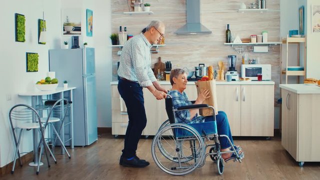 Husband pushing invalid senior woman in wheelchair with grocery paper bag after arriving home from supermarket. Mature people with fresh vegetables for cooking breakfast.