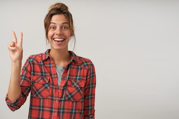 Cheerful young pretty brown haired female with natural makeup forming victory sign while looking gladly at camera with broad smile, isolated over white background