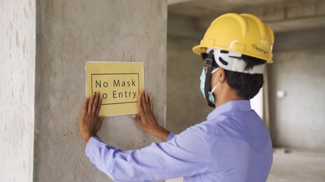 worker pasting No Mask No Entry signage notice borad on wall at working construction site to protect from coronavirus or covid-19 at workplaces - concept of health and labour safety during pandemic.