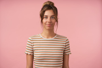 Attractive young green-eyed brunette female smiling gently while looking positively at camera, keeping hands down while standing over pink background