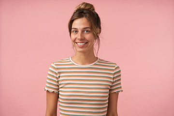 Studio photo of young lovely brown haired lady with bun hairstyle looking at camera with charming smile and keeping hands down while standing over pink background