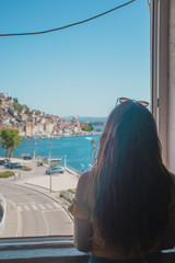 Fototapeta na wymiar Brunette with long hair seen from behind, standing on a window frame looking into the distance. Outline of the town of Sibenik in the distance. Traveling Croatia and the adriatic sea