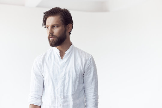 high key portrait photo on white cyclorama of a handsome bearded man with brown hair and eyes, he is wearing a white linen shirt and looks away