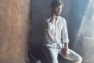 photo of a handsome bearded man with brown hair who sits on a chair in the studio with a ray of sunshine, he is wearing a white linen shirt and pants and looks at the camera