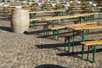 Naklejka premium Wooden tables and benches arranged in rows on the Minster place in Freiburg am Breisgau, Germany. Some of tables are covered with blue and white table cloth. There is a wooden barrel. 