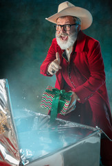 Gifts for 2021. Modern stylish Santa Claus in red fashionable suit and cowboy's hat on dark background. Looks like a rockstar. New Year and Christmas eve, celebration, holidays, winter's mood, fashion