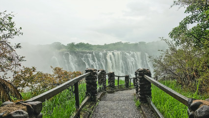 A stone path with railings leads to the unique Victoria Falls. Powerful streams of water fall into...