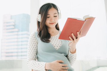 Pregnant asian woman listen to music with wireless headphone and reading a book to tell story to...