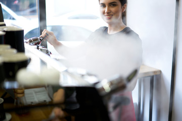 Portrait of cheerful barista enjoying working process of coffee preparation steaming milk for cappuccino.Attentive female employee of cafeteria staff making drinks on professional equipment machine