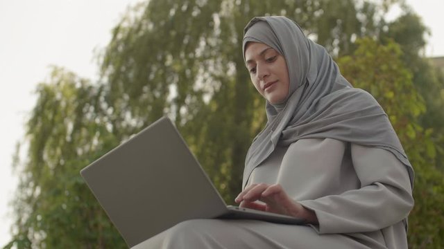 Low angle view of young pretty Muslim woman is sitting outdoors with laptop on her knees, working then looking at camera and smiling