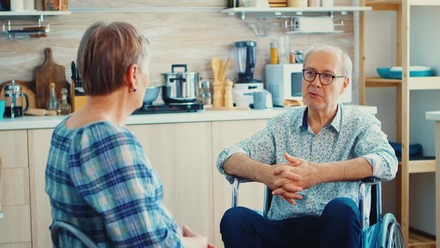 Old woman and her disabled husband in wheelchair chatting in kitchen.Elderly person having a conversation with husband in kitchen. Living with disabled person with walking disabilities