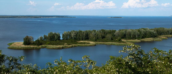 Green oblong island with trees and bushes on a large wide river
