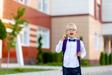 surprised boy schoolboy blond in glasses with a backpack stands at the school and shows a thumbs up. Day of knowledge