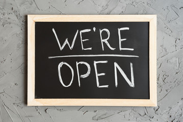 We're Open sign board for cafes and shops. Blackboard with handwritten text, concrete wall as background.