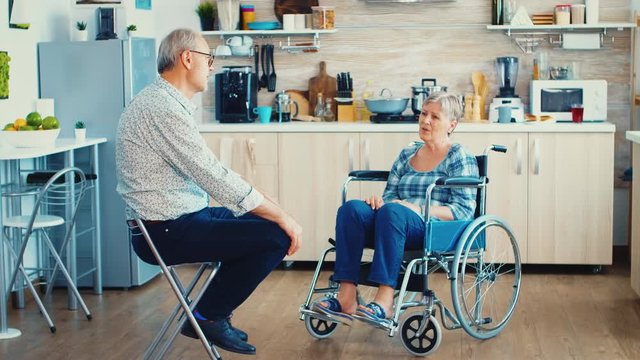 Disabled woman sitting in wheelchair having a conversation with elderly husband in kitchen. Old man having a talking with wife. Living with handicapped person with walking disabilities