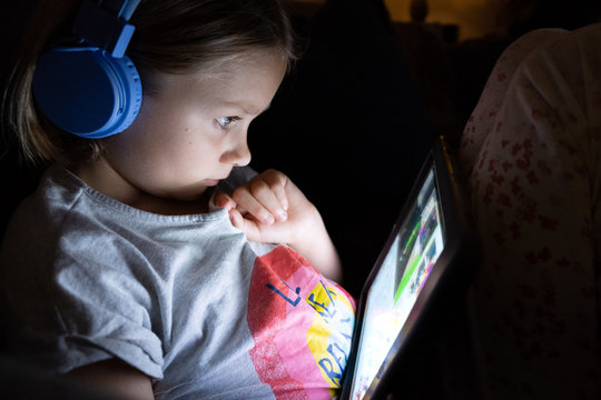 Little Girl On A Tablet With Blue Head Phones In A Dark Room