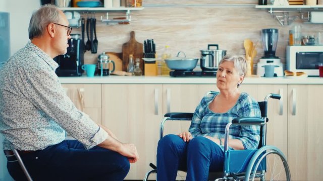 Retired invalid woman in wheelchair having a conversation with old elderly husband in kitchen. Old man talking with wife. Living with disabled person with walking disabilities
