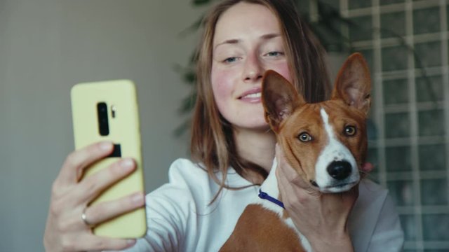 Close-up portrait of beautiful young girl and basenji puppy dog taking selfie at home sitting in living room, looking at camera holding mobile phone. People and animals friendship.