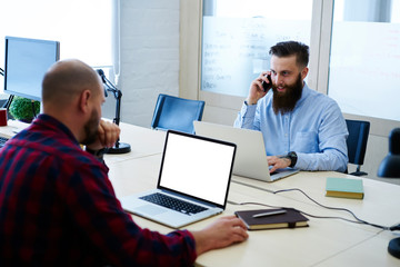 Back view of IT programmer looking at blank screen on laptop with mock up copy space for your advertising content sitting against positive colleague talking on telephone using application in office
