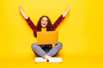 Winner. Excited smiling girl sitting on floor with laptop, raising one hand in the air is she wins, isolated on yellow background