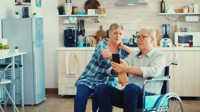 Senior woman and disabled husband in wheelchair surfing on internet using smartphone in kitchen. Paralysied handicapped old elderly man using modern communication techonolgy.