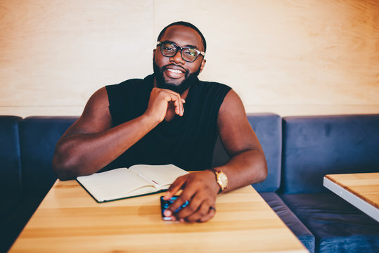 Portrait of afro american muscular guy resting in cafe interior thinking about successful idea for startup, smiling dark-skinned man in eyewear looking at camera enjoying free time planning project