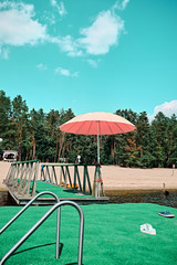 There is a pink beach umbrella on the green pier. Sunny weekend. Pines can be seen on the river bank.