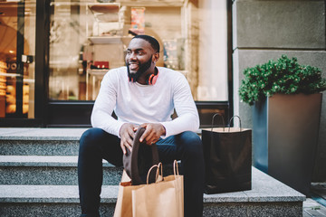 Smiling african american male customer in trendy wear sitting on stairs of store with bags with...