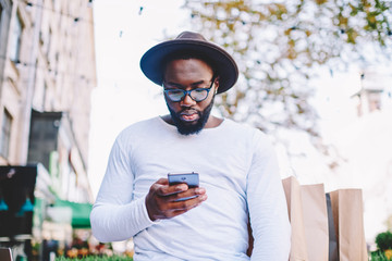 Pensive african american hipster guy in trendy hat installing application on smartphone spending time outdoors,thoughtful dark skinned man in shirt with copy space for brand name reading news on phone