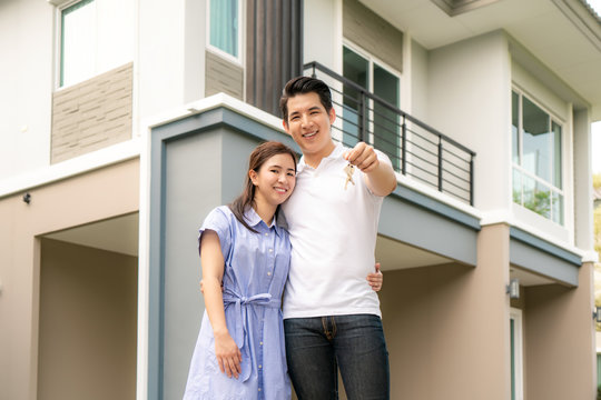 Portrait of Asian young couple standing and hugging together and holding house key looking happy in front of their new house to start new life. Family, age, home, real estate and people concept..