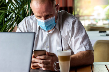 Fototapeta na wymiar Businessman with face mask sitting in a restaurant looking at mobile while working on a laptop. Photo of businessman working in cafe,drinking coffee and browsing internet using smartphone during break