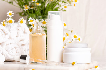 Glass and plastic bottles of essence or serum and with chamomile flowers on marble board. Moisturizing face cream and camomile