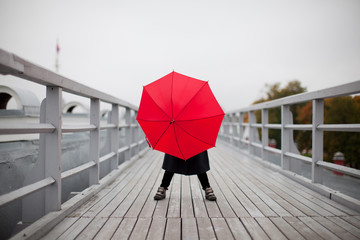 a child in autumn clothes hiding behind red umbrella on a cloudy and rainy day.