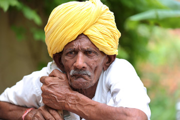 Amazed old Indian man wearing yellow turban looking up with mouth opened. Curious old man. Wearing traditional Indian clothes. Old man holding stick in hand. Old man waiting for his turn. 