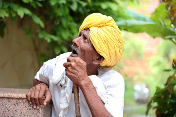 Amazed old Indian man wearing yellow turban looking up with mouth opened. Curious old man. Wearing traditional Indian clothes. Old man holding stick in hand. Old man waiting for his turn. 