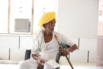 Amazed old Indian man wearing yellow turban looking up with mouth opened. Curious old man. Wearing traditional Indian clothes. Old man holding stick in hand. Old man waiting for his turn.