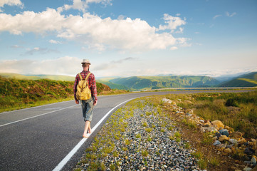 A young bearded hipster in a shirt hat and barefoot walks along a country road in the mountains. Hitchhiking and free travel