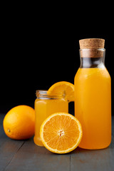fresh orange juice in a glass jar and bottle with fresh fruits on wooden table, black background