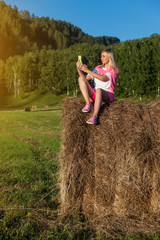 Naklejka na ściany i meble Blonde girl in bright pink suit and white T-shirt sits on dry haystack against background of mountains, trees and round rolls of hay with phone and takes selfie with hand outstretched. Summer vacation