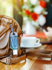 Mini alcohol gel bottle to kill Corona Virus(Covid-19) hang on a brown leather shoulder bag with hot coffee on table in cafe.New normal lifestyle.Health care concept.Selective focus on alcohol gel