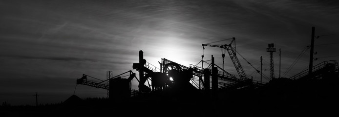 Silhouettes of constructions and equipment in a mining enterprise against the backdrop of a sunset, black and white panorama. Industrial landscape.