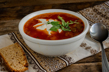 Borscht in a white bowl, bread, metal spoon, kitchen folk. Napkin in the home kitchen on a wooden table.