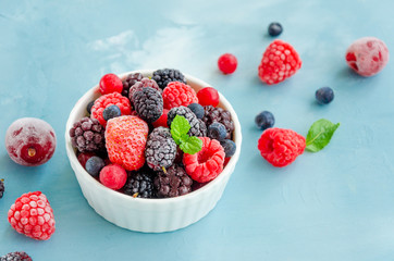 Mix of summer frozen berries in a white bowl on a blue concrete background. Copy space.