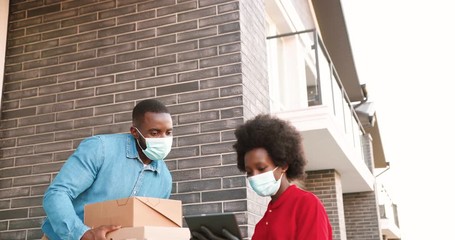 African American female courier in mask delivering carton parcels to male client at house and handing outside. Delivery woman bringing pizza boxes to man customer. Coronavirus concept.