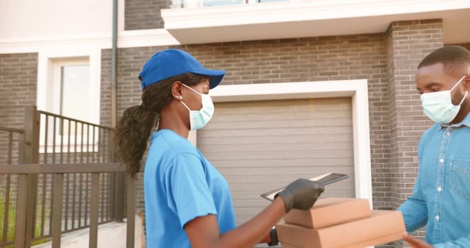 African American female courier in medical mask delivering carton parcels to male client at house and handing outdoors. Delivery woman bringing boxes to man and using tablet device Pandemic quarantine
