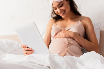 selective focus of pregnant woman in silk nightie holding digital tablet and touching belly in bedroom