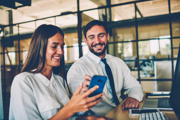 Cheerful female employee showing funny video on smartphone to her colleague during work break,coworkers having fun together in office reading news from networks and joking enjoying friendly talk