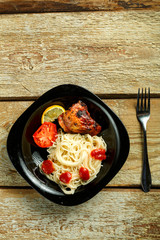 Black plate of spaghetti with baked chicken near tomatoes and garlic on a wooden table.